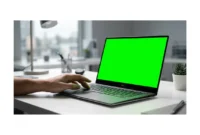 Modern laptop displaying a green screen tint on a clean desk with a user's hand reaching toward the keyboard, representing common laptop green screen issues and troubleshooting solutions