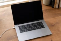 MacBook laptop open on a desk with a black screen and charging cable connected, showing a MacBook that won't turn on but is charging CHARACTERS: 136 (trim to: MacBook open on desk with black screen and charging cable plugged in, not turning on while charging)