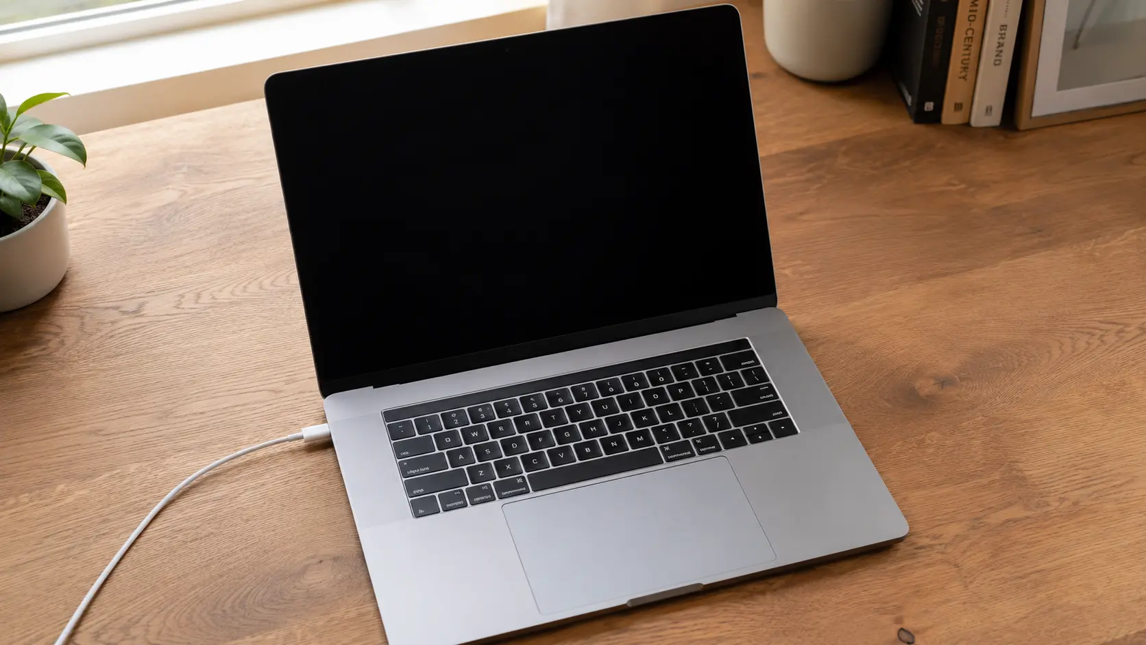 MacBook laptop open on a desk with a black screen and charging cable connected, showing a MacBook that won't turn on but is charging CHARACTERS: 136 (trim to: MacBook open on desk with black screen and charging cable plugged in, not turning on while charging)
